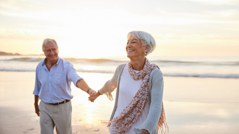 A senior man and a senior woman walking on a beach and holding hands.