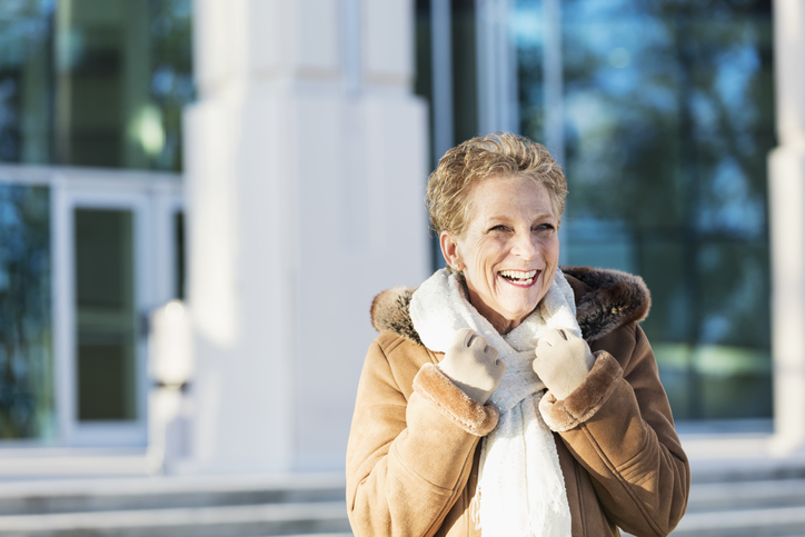 Senior woman in winter coat standing outside building in the sun.