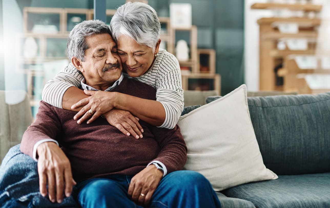 Elderly woman hugging her husband who's on the couch at home.